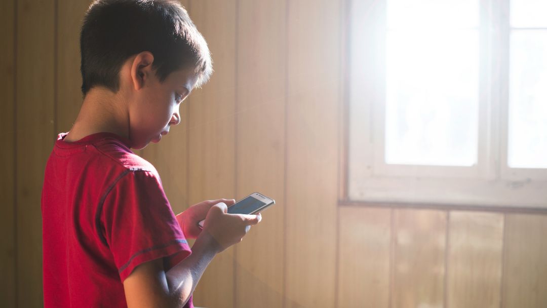 Young child standing alone by a window looking at a phone, representing early exposure to harmful content