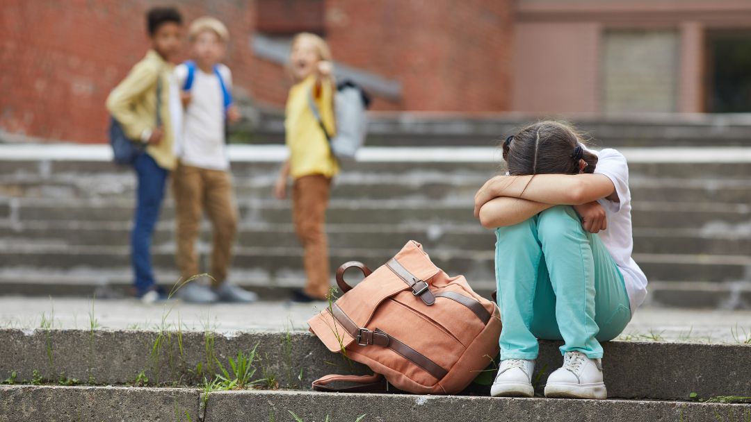Young girl sitting alone outside school, excluded, representing the everyday harassment girls experience